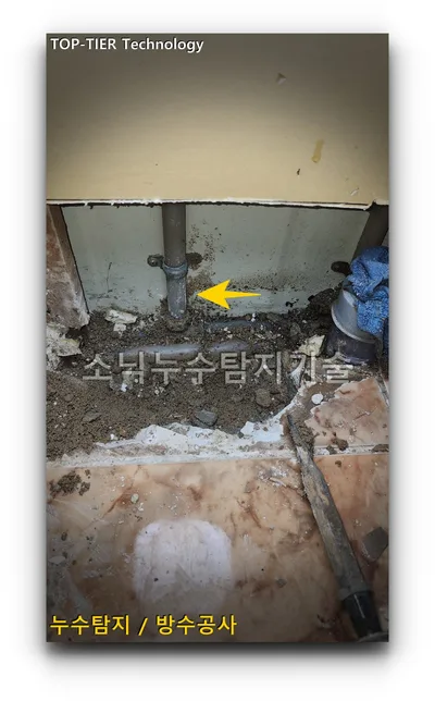 타일을 걷어낸 싱크대 아래 바닥에서 회색 배관이 드러나고 노란 화살표가 배관 아래 지점을 가리키는 모습